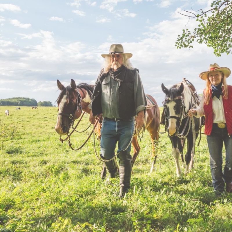 Mann und Frau in Cowboy-Kluft spazieren mit zwei Pferden über Wiese