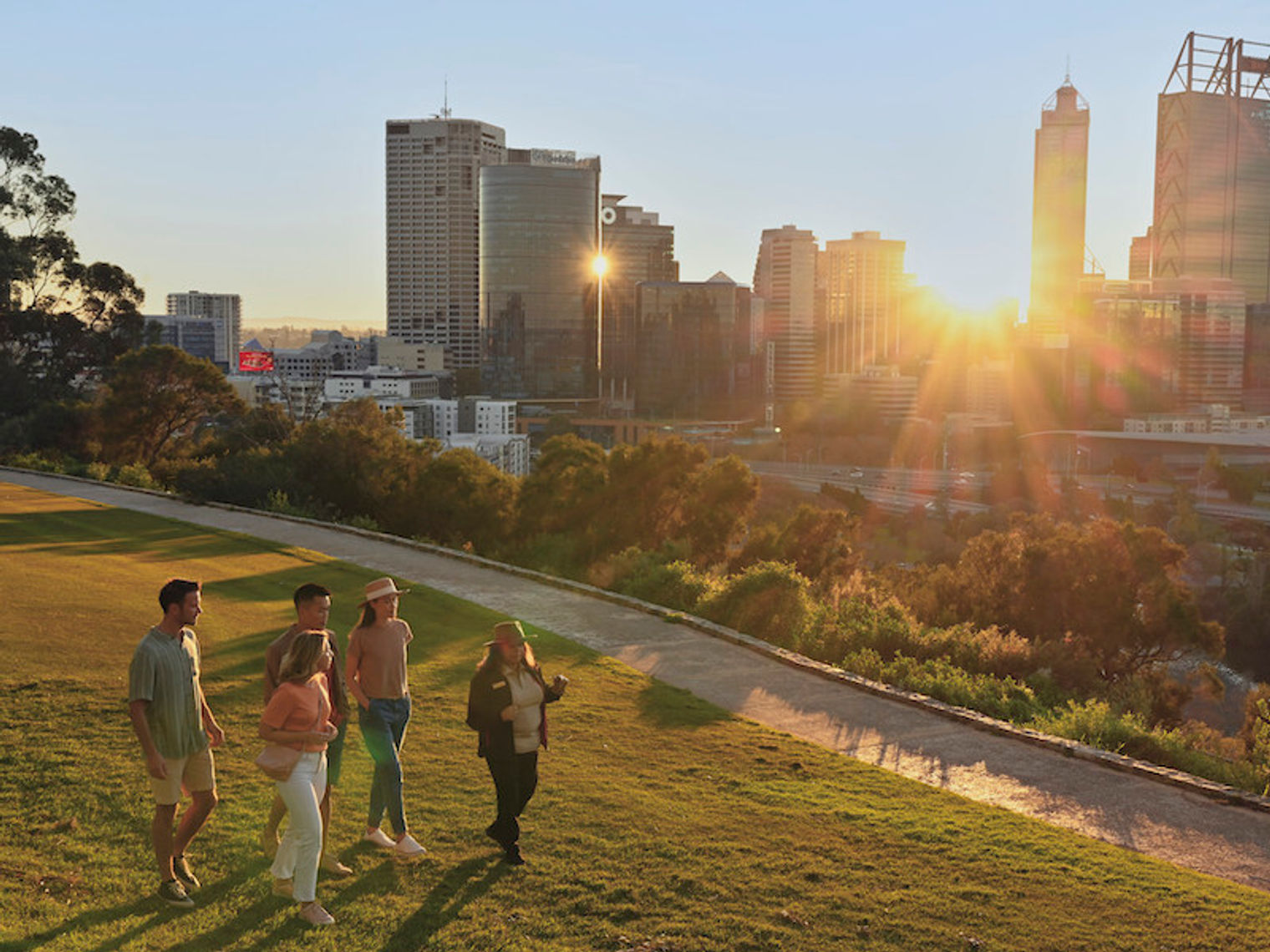 Eine Gruppe von Menschen, die im King's Park in Perth spazieren gehen, während die Sonne zwischen den Gebäuden der Stadt im Hintergrund scheint, in Australien