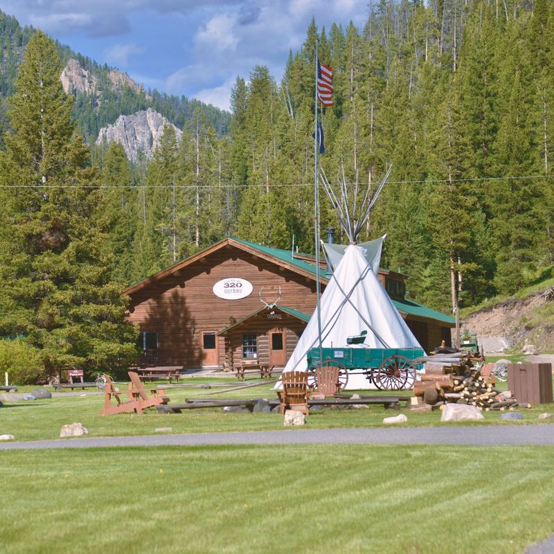 Het hoofdgebouw van 320 Guest Ranch met een grote tipi, een vintage wagen, Adirondack-stoelen en blokhutjes op een groen gazon, omgeven door dennenbos en rotsachtige bergtoppen bij West Yellowstone, Montana, VS.