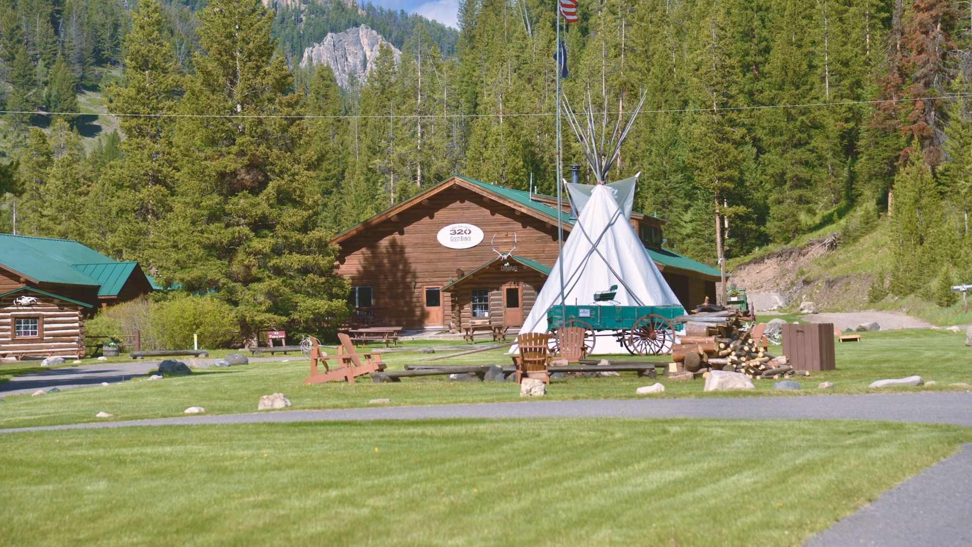 The main lodge of 320 Guest Ranch with a large tipi, vintage wagon, Adirondack chairs, and log cabins on a green lawn, surrounded by pine forest and rocky mountain peaks near West Yellowstone, Montana, USA.