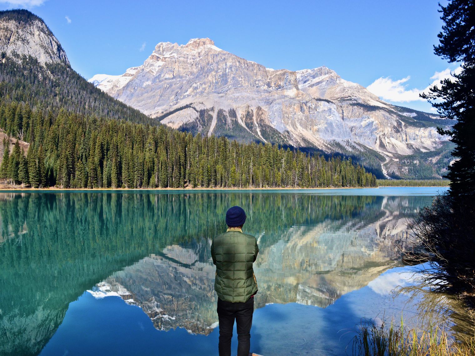 Eine Person steht an einem türkisfarbenen See in Kanada und blickt auf das bewaldete Ufer und die hoch aufragenden Gipfel der Rocky Mountains, die sich im stillen Wasser spiegeln.