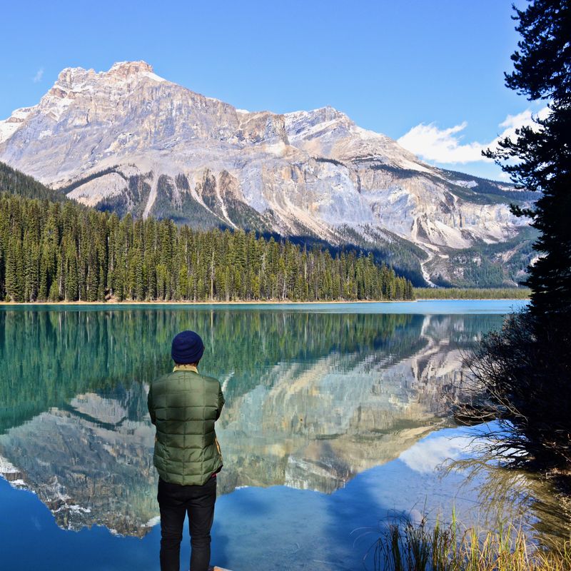 Eine Person steht an einem türkisfarbenen See in Kanada und blickt auf das bewaldete Ufer und die hoch aufragenden Gipfel der Rocky Mountains, die sich im stillen Wasser spiegeln.