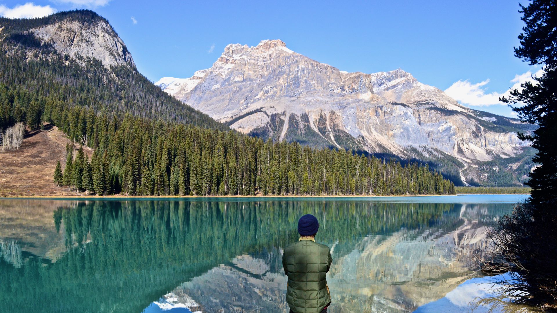 Een persoon die bij een turquoise meer in Canada staat en uitkijkt over de beboste kustlijn en de torenhoge toppen van de Rocky Mountains die weerspiegeld worden in het stille water.