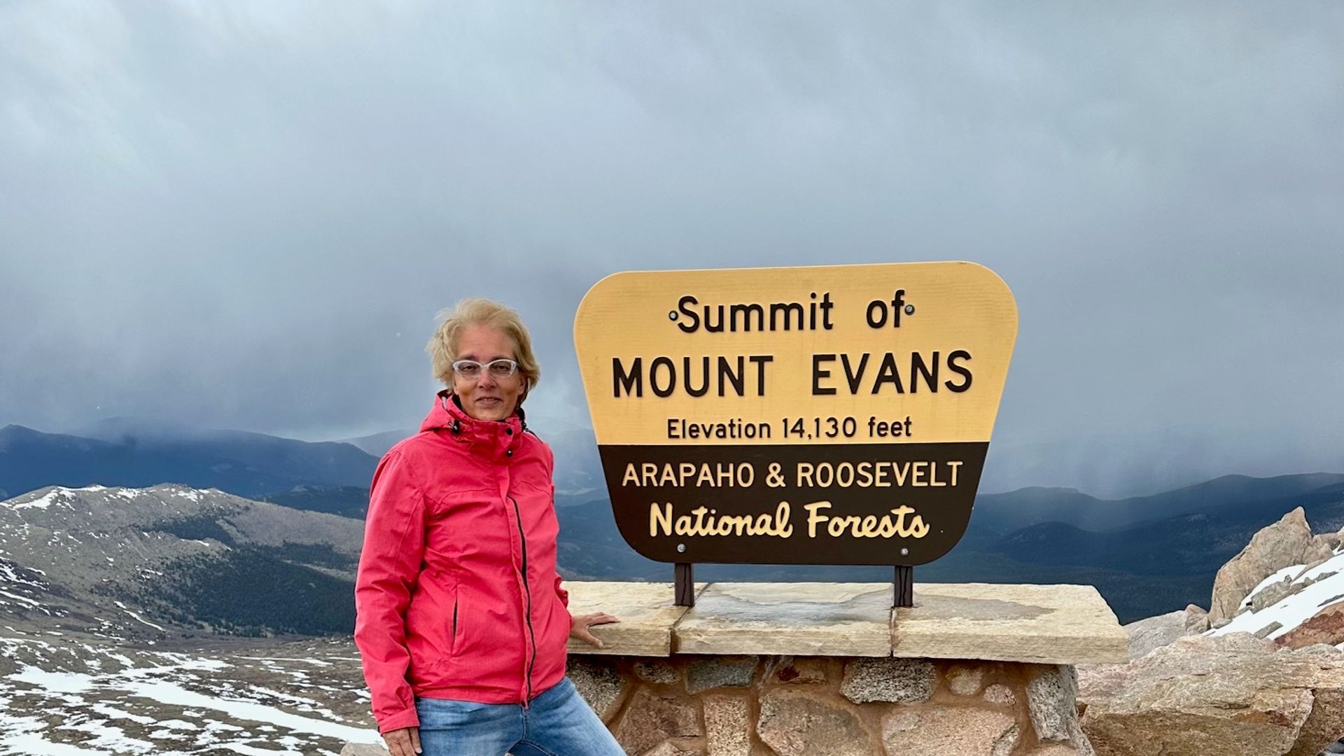 Mevrouw Cesar staat bij het summit bord op de top van Mount Evans