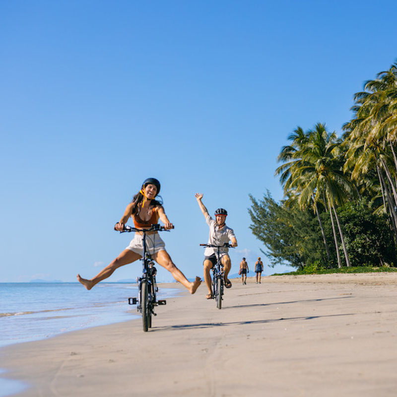 Een vrolijk stel fietst over het strand terwijl de man een hand in de lucht steekt en de vrouw haar voeten van de pedalen houdt