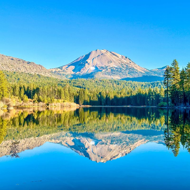 Een rustig meer in Californië dat de omringende dennenbossen weerspiegelt en de opvallende vulkanische piek die in de verte oprijst onder een helderblauwe hemel.