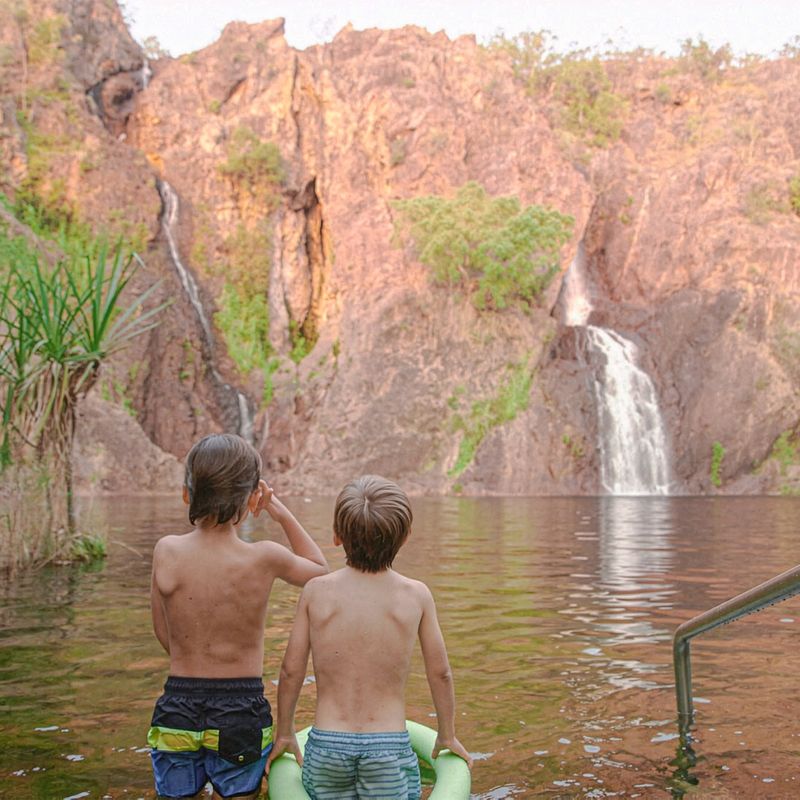 Zwei Kinder waten in einem natürlichen Felsenbecken und blicken auf einen Wasserfall, der an den roten Sandsteinfelsen der Wangi Falls im Litchfield-Nationalpark im Northern Territory, Australien, herabstürzt