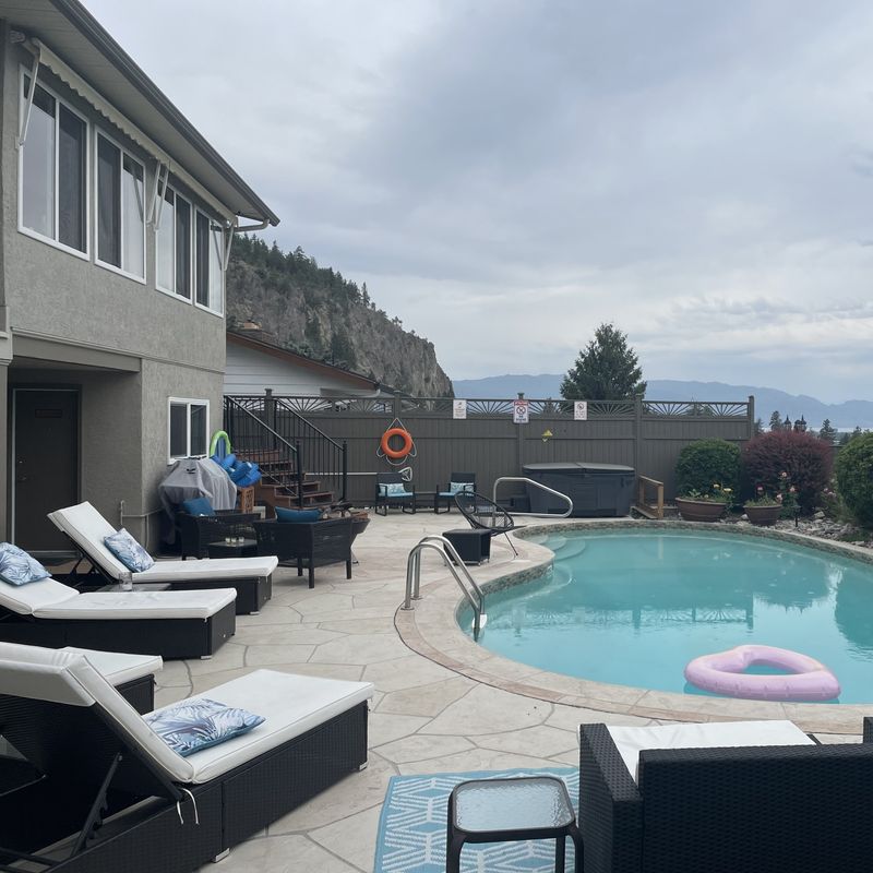 Een privé buitenzwembad met roze opblaasbanden en ligbedden op een betegeld terras, met een jacuzzi, tuinstoelen en uitzicht op het Okanagan Lake en de omliggende bergen achter het hek, bij een villa in de Okanagan Valley, Brits-Columbia, Canada.