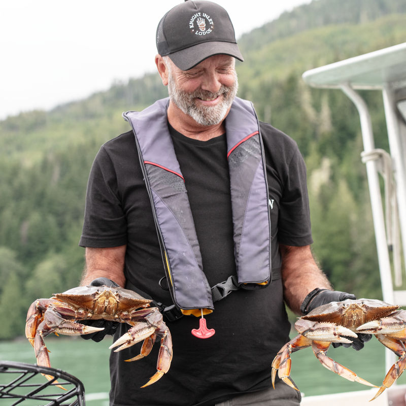 Ein lächelnder Guide der Knight Inlet Lodge hält zwei frisch gefangene Dungeness-Krabben auf einem Boot in die Höhe, mit bewaldeten Fjordhängen im Hintergrund, am Knight Inlet, British Columbia, Kanada.