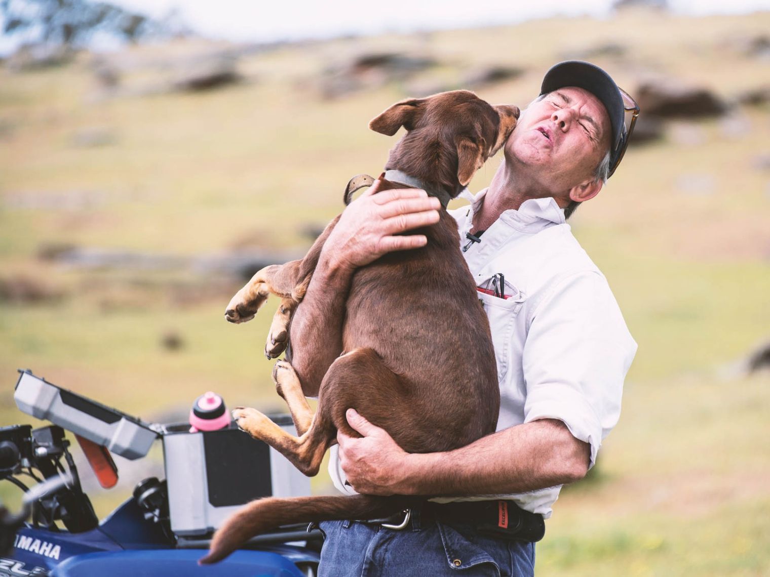 A man holds his dog who's trying to lick him on the face with a quad bike next to them