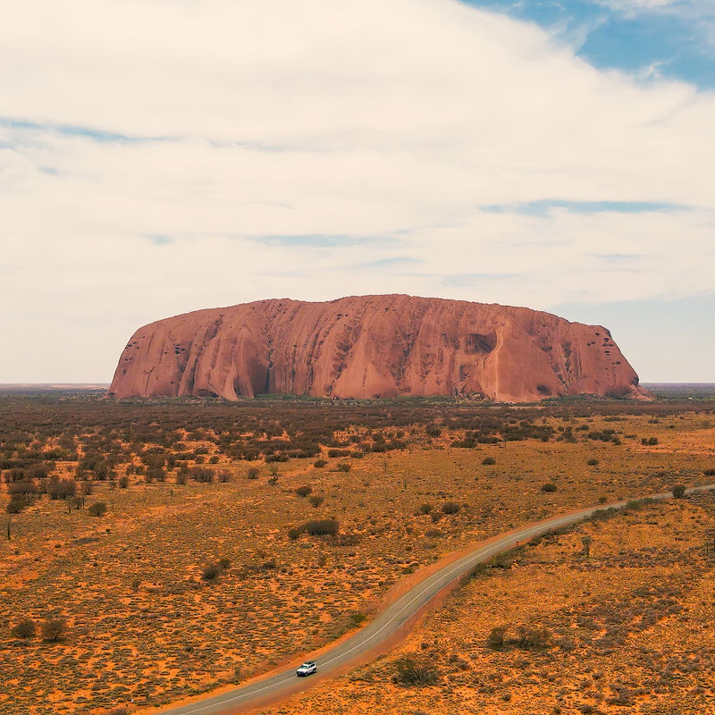 Uluru am Horizont, ein Auto fährt davor auf einer Straße entlang