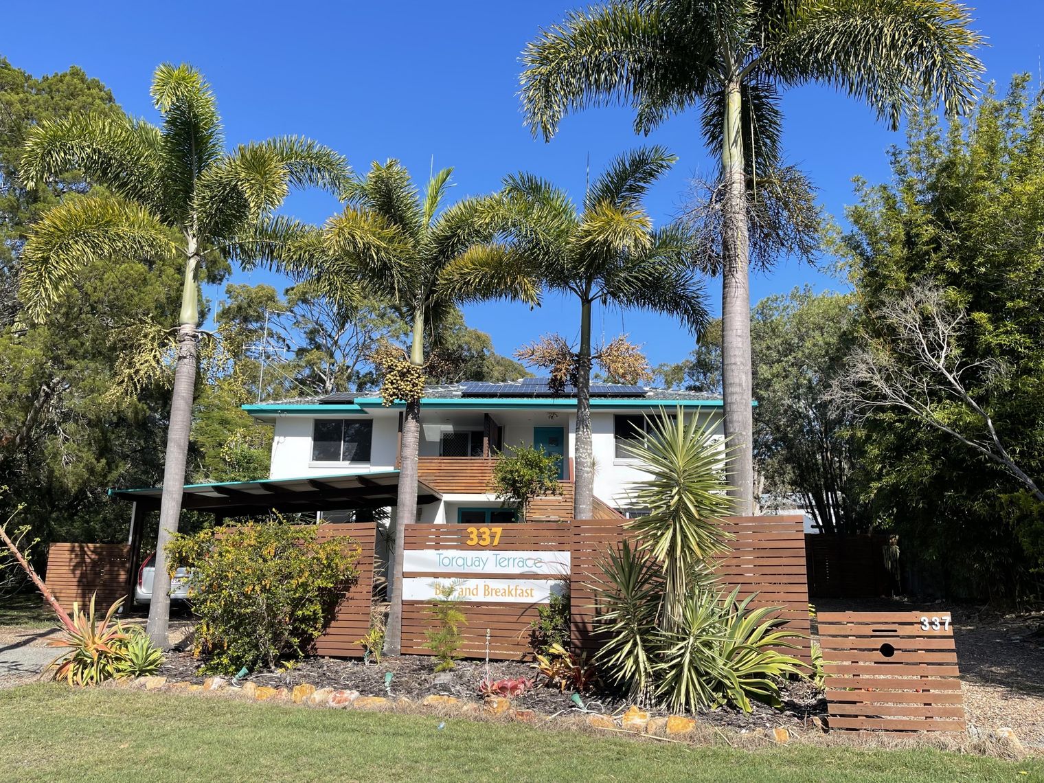 Twee verdiepingen tellende bed & breakfast-accommodatie in Australië, omgeven door hoge palmbomen en tropische tuinen achter een houten schutting, onder een helderblauwe hemel.