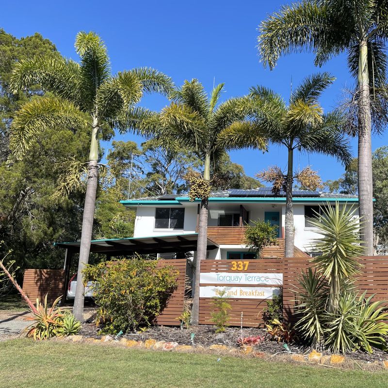 Twee verdiepingen tellende bed & breakfast-accommodatie in Australië, omgeven door hoge palmbomen en tropische tuinen achter een houten schutting, onder een helderblauwe hemel.
