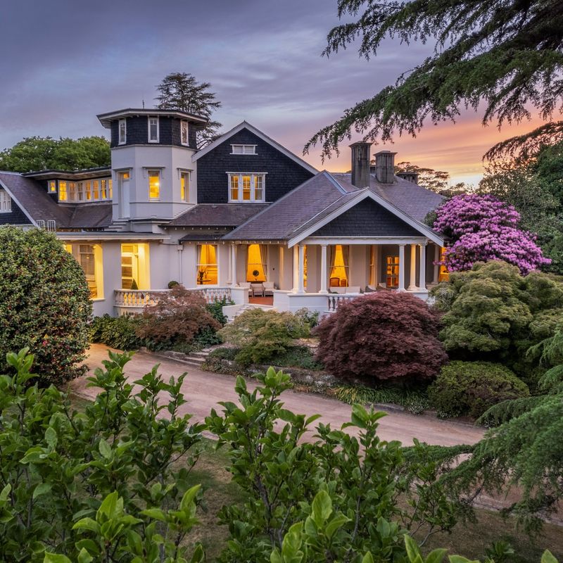 Grote historische bed & breakfast-accommodatie in Australië, met een monumentaal landhuis met rondomlopende veranda en toren, omgeven door verzorgde tuinen met Japanse esdoorns en bloeiende rododendrons bij zonsondergang.