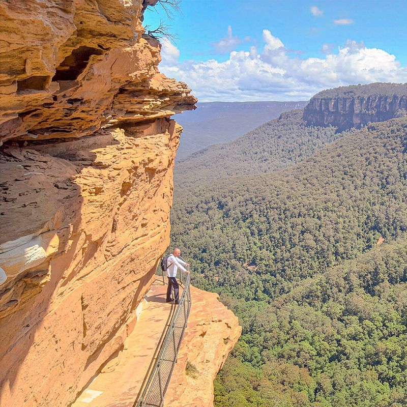Een wandelaar staat op een wandelpad langs de rotswand en kijkt uit over de uitgestrekte beboste valleien en zandstenen rotswanden van de Blue Mountains, New South Wales, Australië