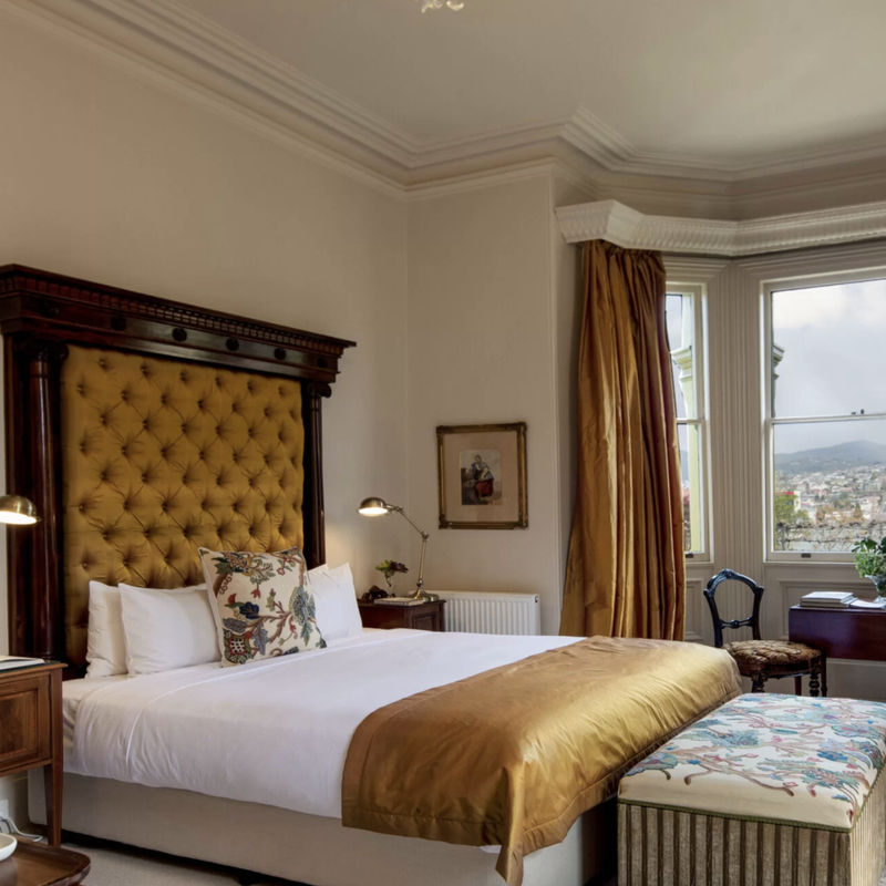 Bedroom with a large upholstered headboard, gold curtains, a window overlooking a hillside town, and antique-style furniture.