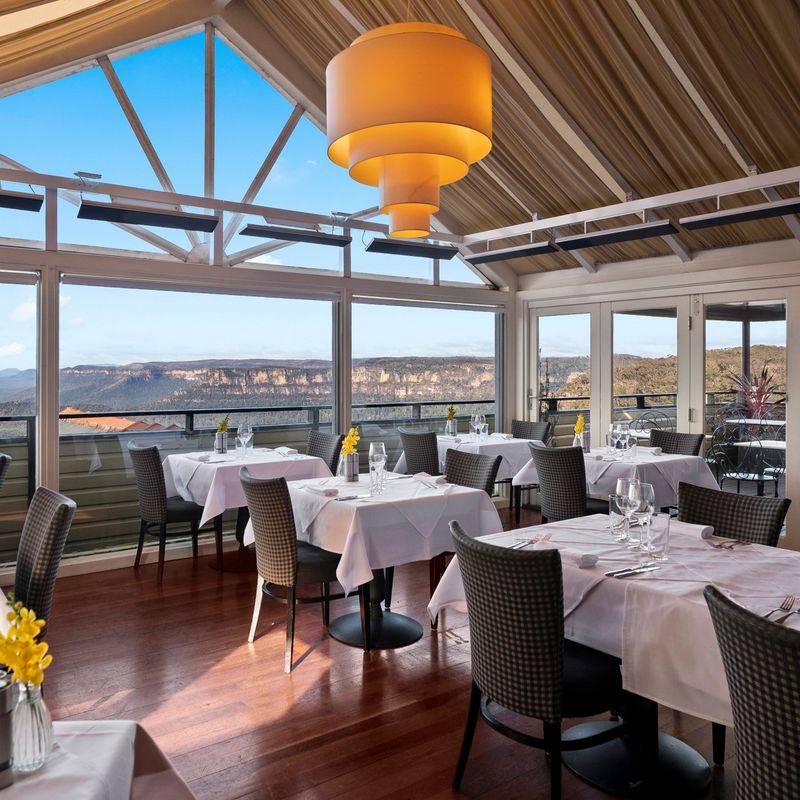 Stijlvol boetiekhotel-restaurant in Australië met witgedekte eettafels en panoramisch uitzicht over de Blue Mountains door kamerhoge ramen onder een gewelfd houten plafond en bergen op de achtergrond.