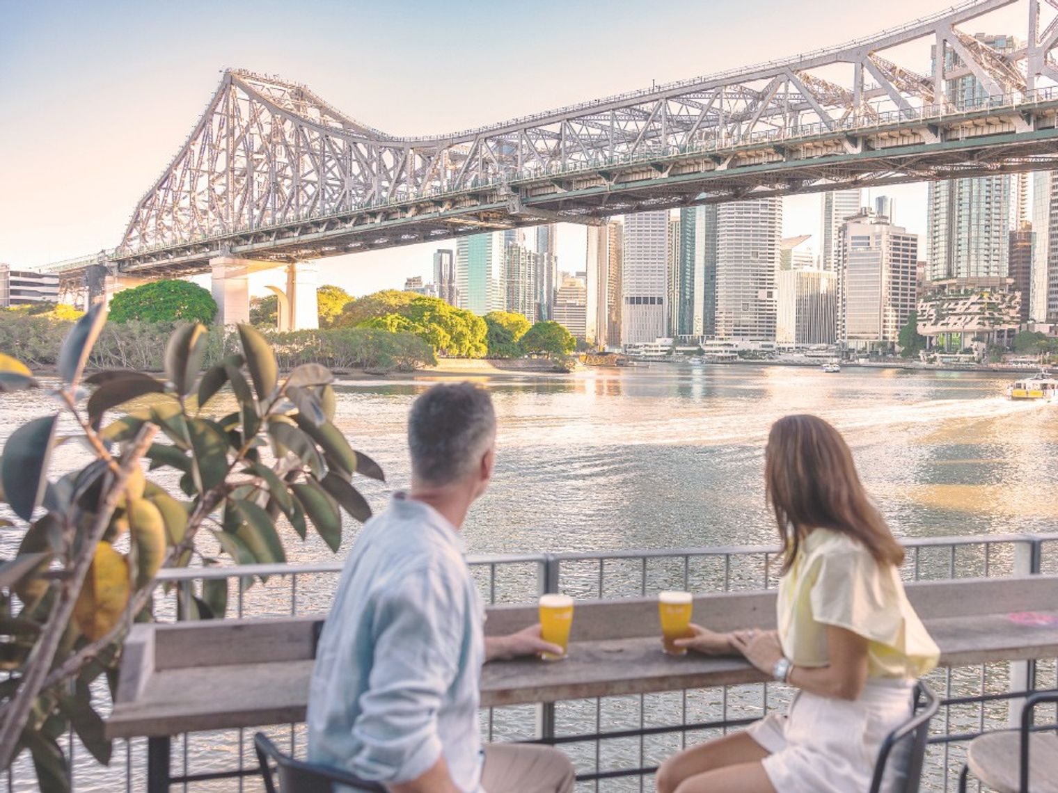 Two people sit at a table with glasses of beer looking out at the river, bridge and buildings of Brisbane