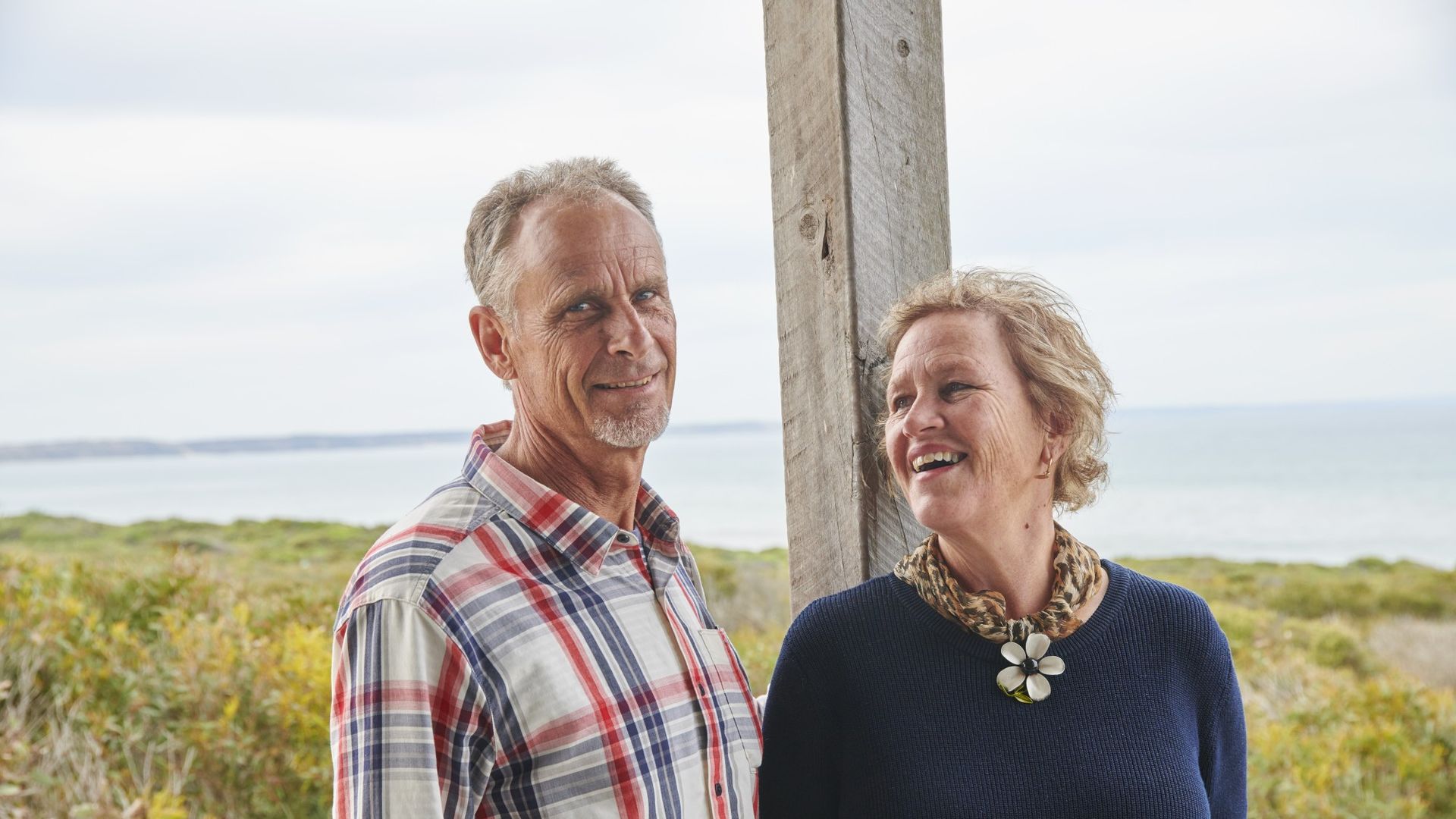 A couple stand against a wooden beam smiling with bush and ocean in the background