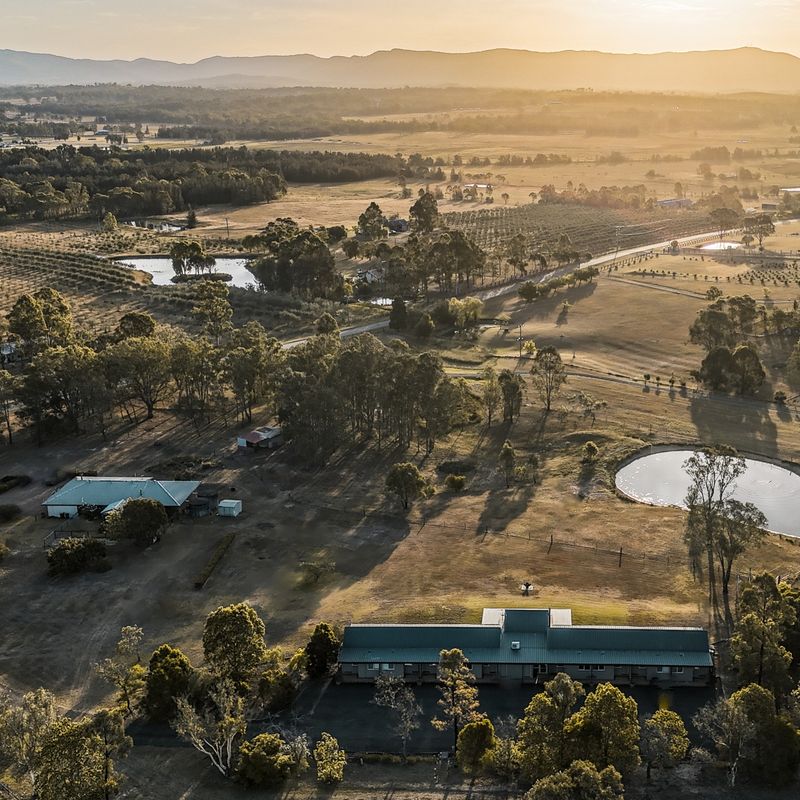 Luchtfoto van een boerderijaccommodatie in Australië tijdens het gouden uur, met een landelijke lodge omgeven door glooiende weilanden, boerderijdammen, boomgaarden en eucalyptusbomen met bergen in de mistige achtergrond.
