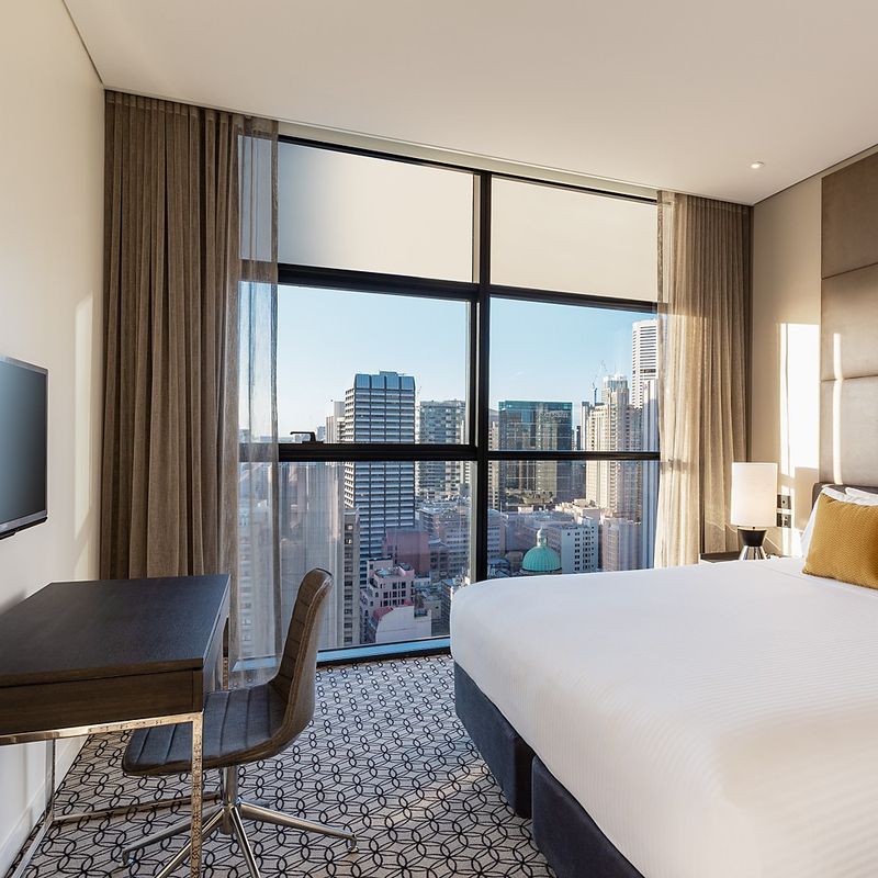 An apartment bedroom with a double bed and desk and chair with windows overlooking the Sydney skyline