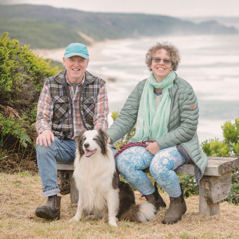 Local hosts Mark and Roz sit on a bench with their dog Blue, with the coastline of the Great Ocean Road in the background