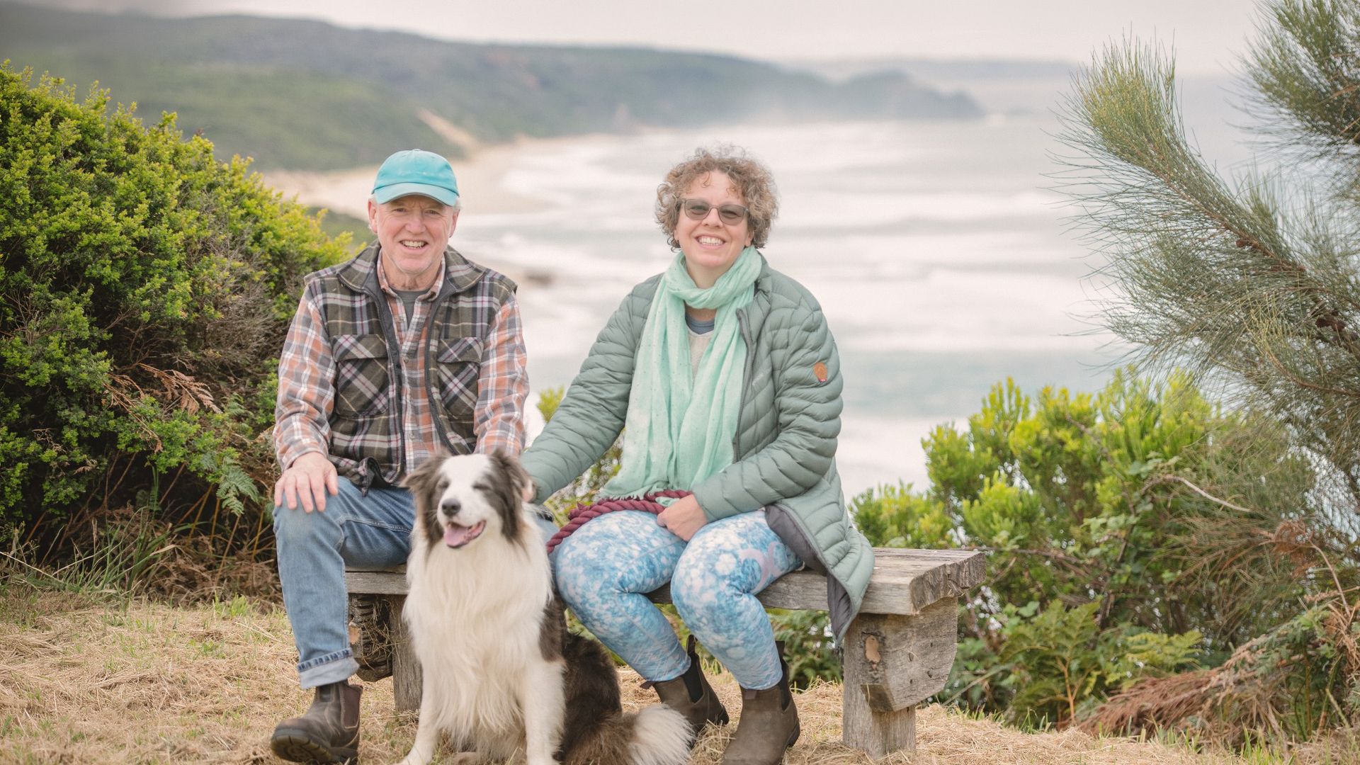 Die einheimischen Gastgeber Mark und Roz sitzen mit ihrem Hund Blue auf einer Bank, im Hintergrund ist die Küste der Great Ocean Road zu sehen