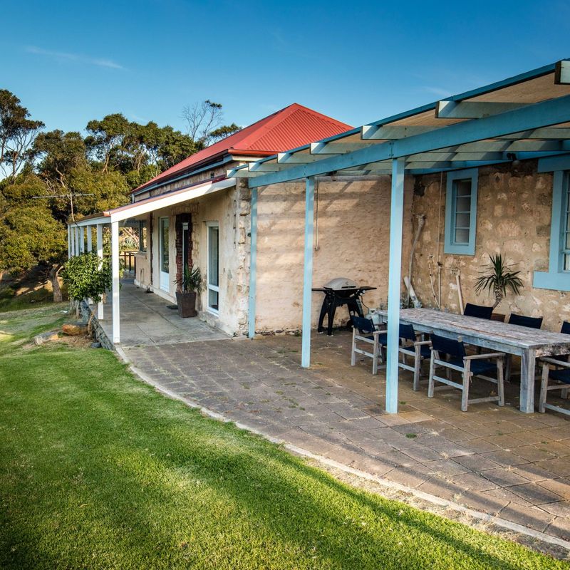 Historisch stenen vakantiehuis in Australië met een overdekt terras om buiten te eten en een barbecue, gelegen op een klif met een gazon en een prachtig uitzicht over de oceaan op een heldere, zonnige dag.