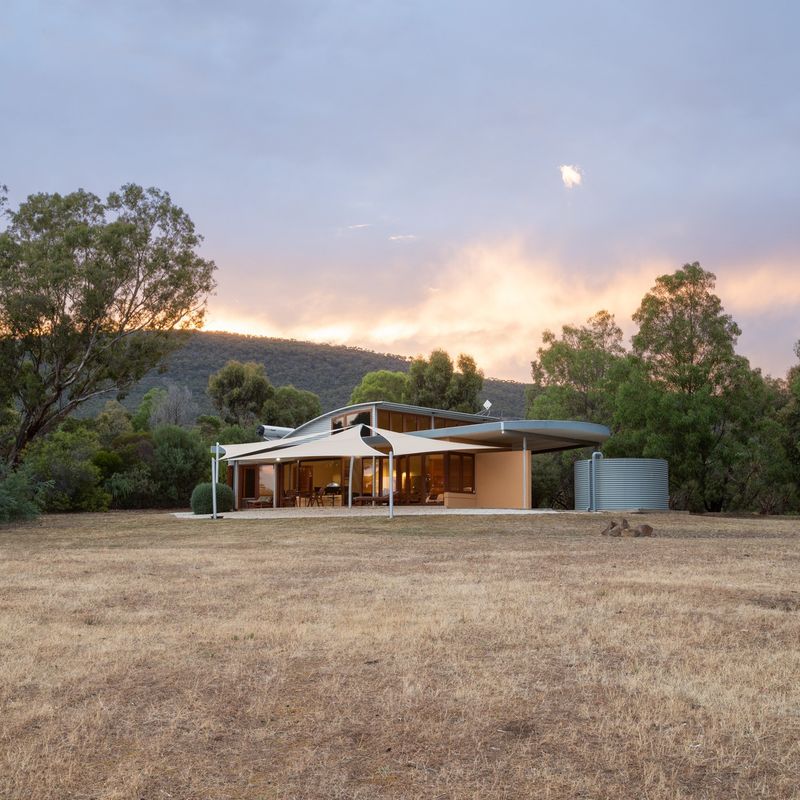 Contemporary architect-designed holiday house with curved roof and sail shades, set in an open dry grassland clearing surrounded by eucalyptus trees and hills at sunset in rural Australia