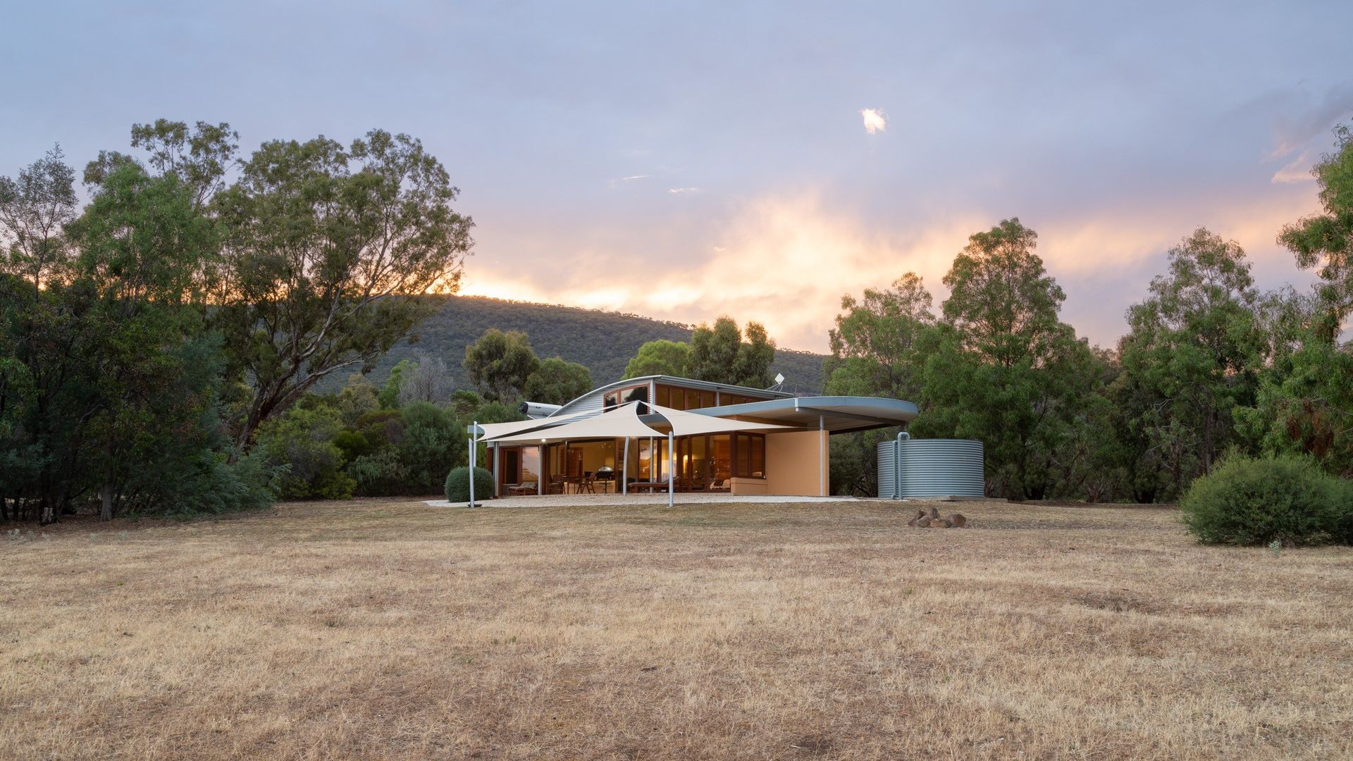 Moderne, door een architect ontworpen vakantiewoning met gebogen dak en zeilzonwering, gelegen in een open, droge graslandweide omringd door eucalyptusbomen en heuvels bij zonsondergang op het Australische platteland.