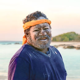 Ein lächelnder australischer Ureinwohner mit traditioneller weißer Gesichtsbemalung und einem orangefarbenen Stirnband an einem tropischen Strand bei Sonnenuntergang, Australien