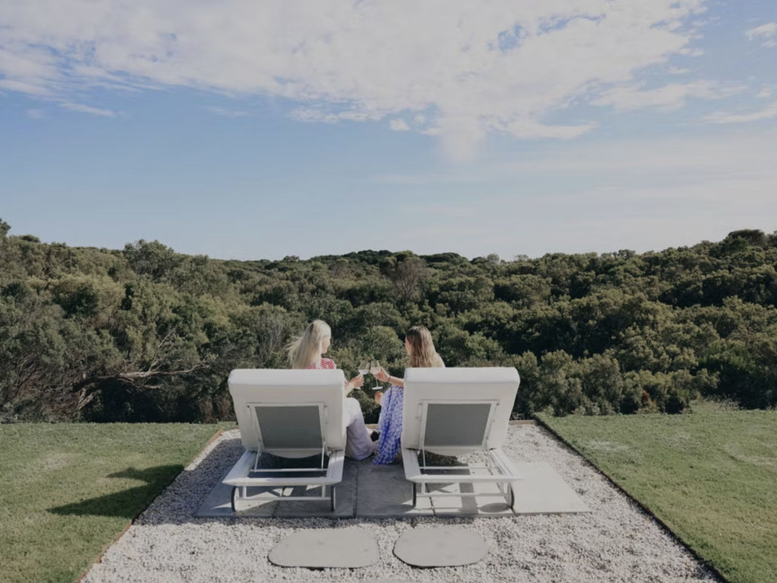 Zwei Personen sitzen auf Liegestühlen im Freien mit Blick auf dichtes Grün und den Himmel und stoßen mit Gläsern an.
