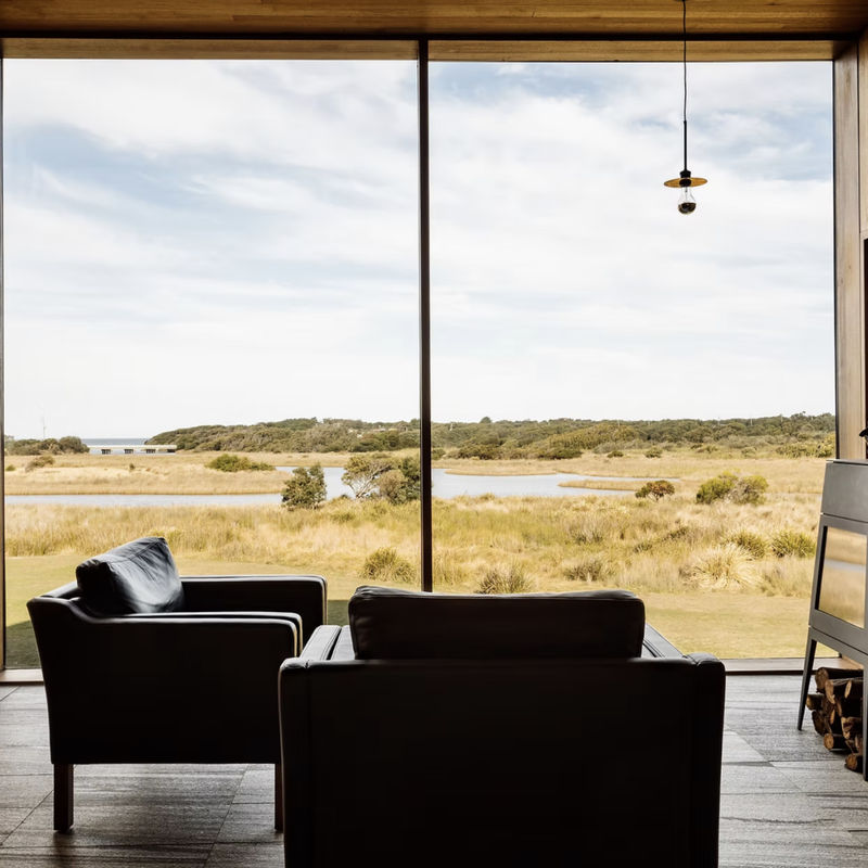 Two armchairs facing a large picture window overlooking open grassland, water and distant bushland, with a wood stove on the right.