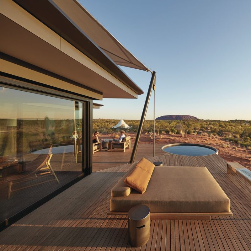 Luxe lodgeterras met ligbed en dompelbad met uitzicht op het rode woestijnlandschap van Uluru in het Northern Territory van Australië, met een stel op de achtergrond tijdens het gouden uur.