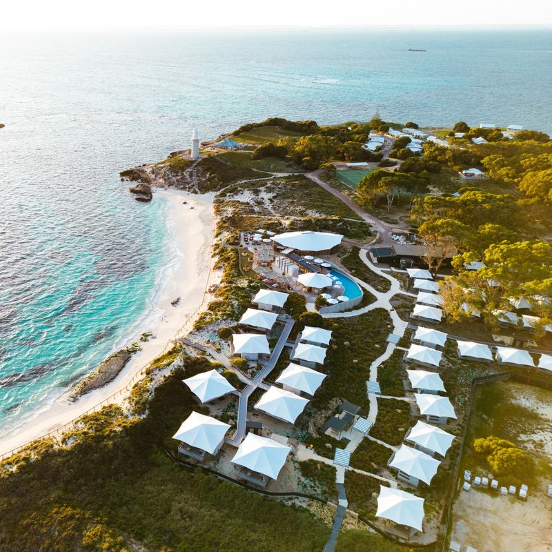 Luchtfoto van een resort aan het strand op Rottnest Island, Australië, met glamping-tenten met witte daken langs een turquoise kustlijn met witte zandstranden en een vuurtoren bij zonsondergang.