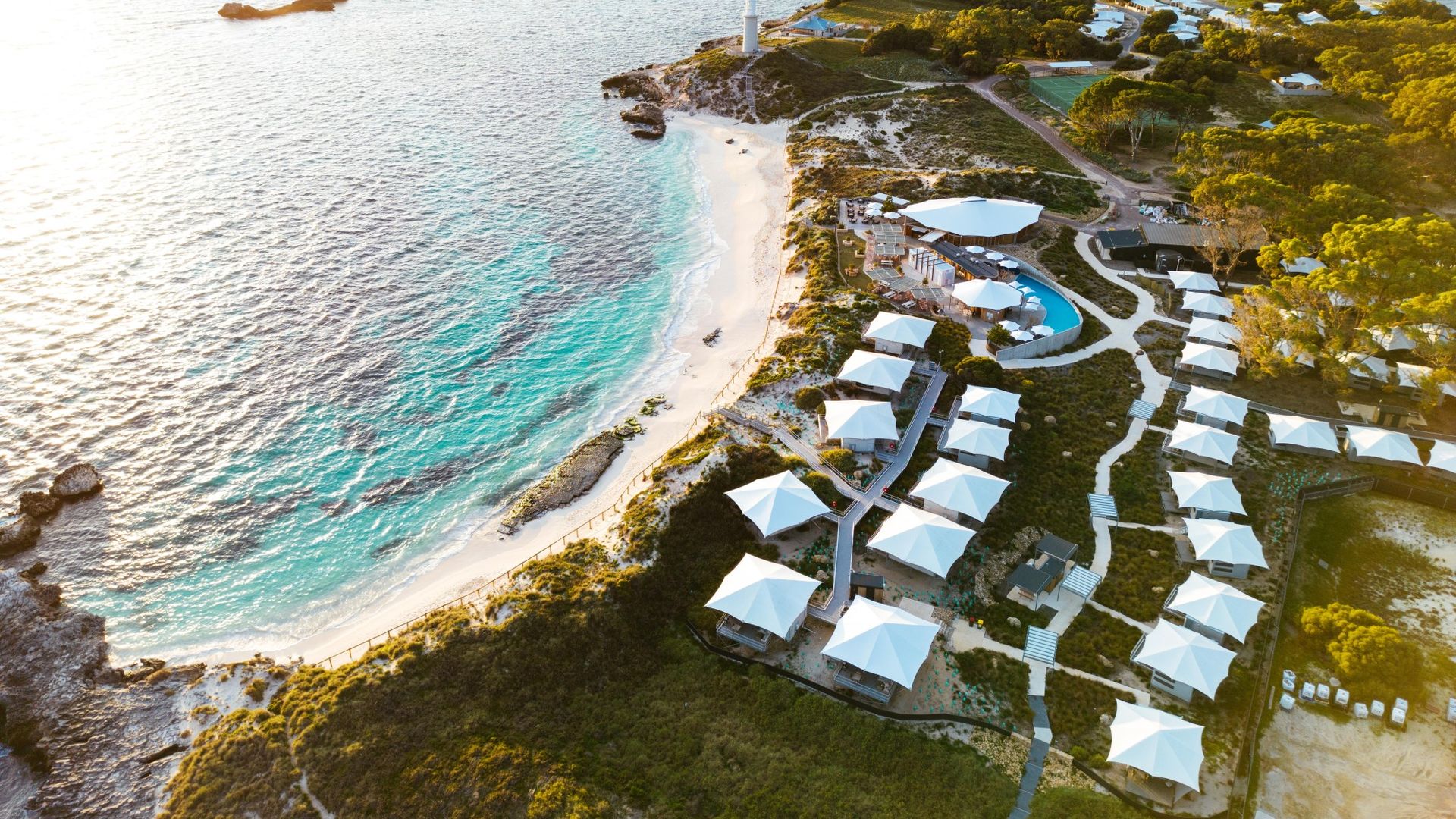 Luftaufnahme eines Strandresorts auf Rottnest Island, Australien, mit weißen Glamping-Zelten entlang einer türkisfarbenen Küste mit weißen Sandstränden und einem Leuchtturm bei Sonnenuntergang