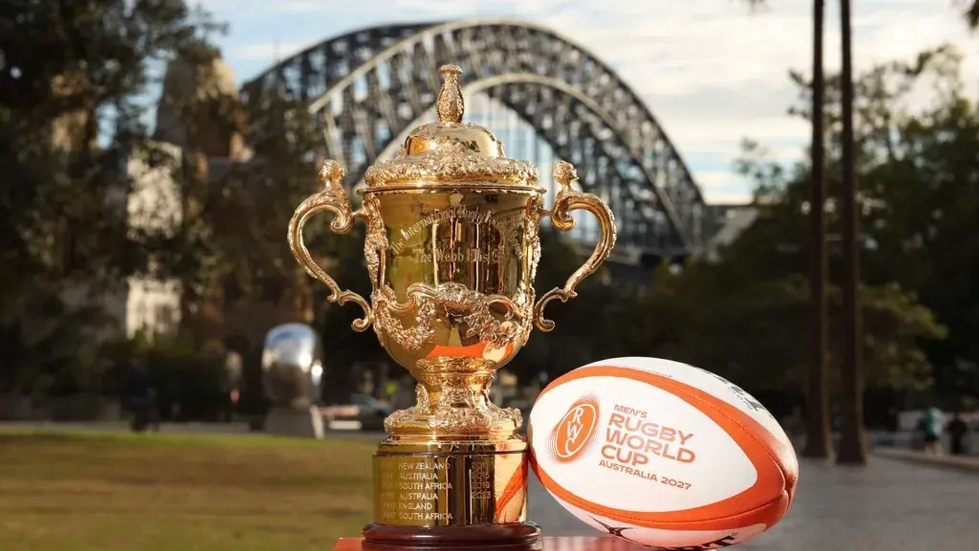 A rugby ball and the Webb Ellis trophy sit on a plinth with the Sydney Harbour Bridge in the background