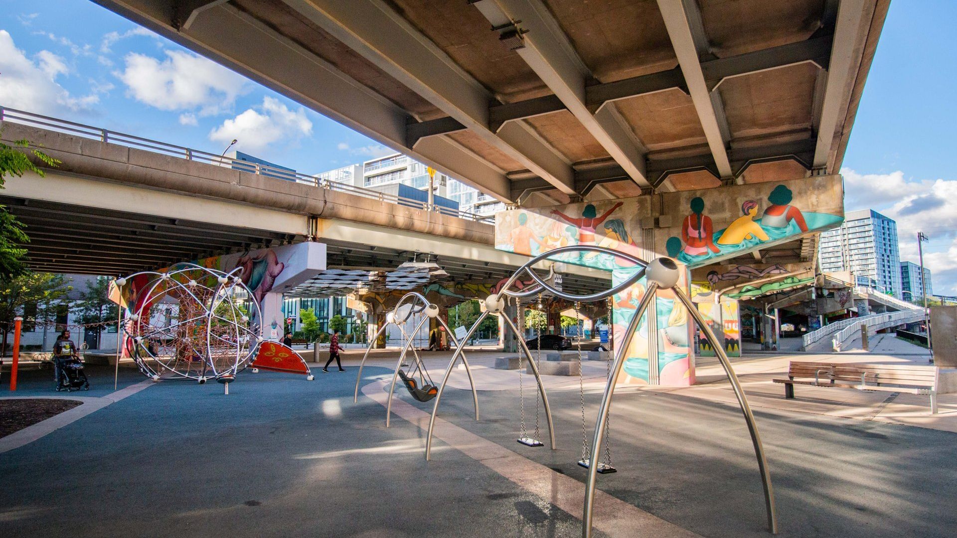Interactive spider-like playground sculpture under a highway overpass with murals in a Toronto urban park