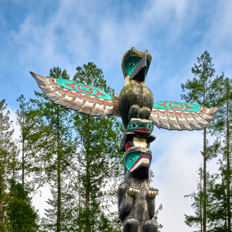 Traditioneller Totempfahl der First Nations mit einem Donnervogel und geschnitzten Figuren, die sich vor einem blauen Himmel über einem Kiefernwald in British Columbia, Kanada, erheben