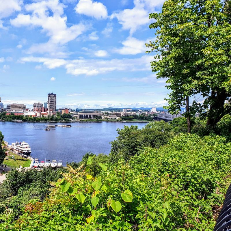 Panoramablick über den Ottawa River auf die Skyline, eingerahmt von üppigem Grün und einem sonnigen Himmel