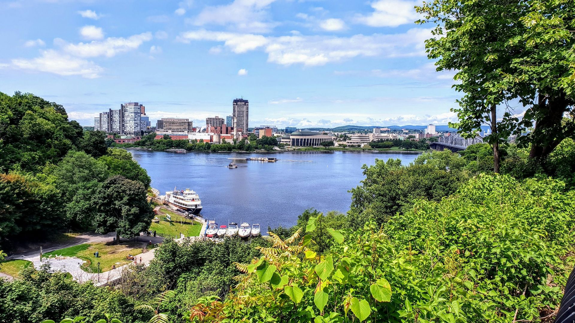 Panoramablick über den Ottawa River auf die Skyline, eingerahmt von üppigem Grün und einem sonnigen Himmel