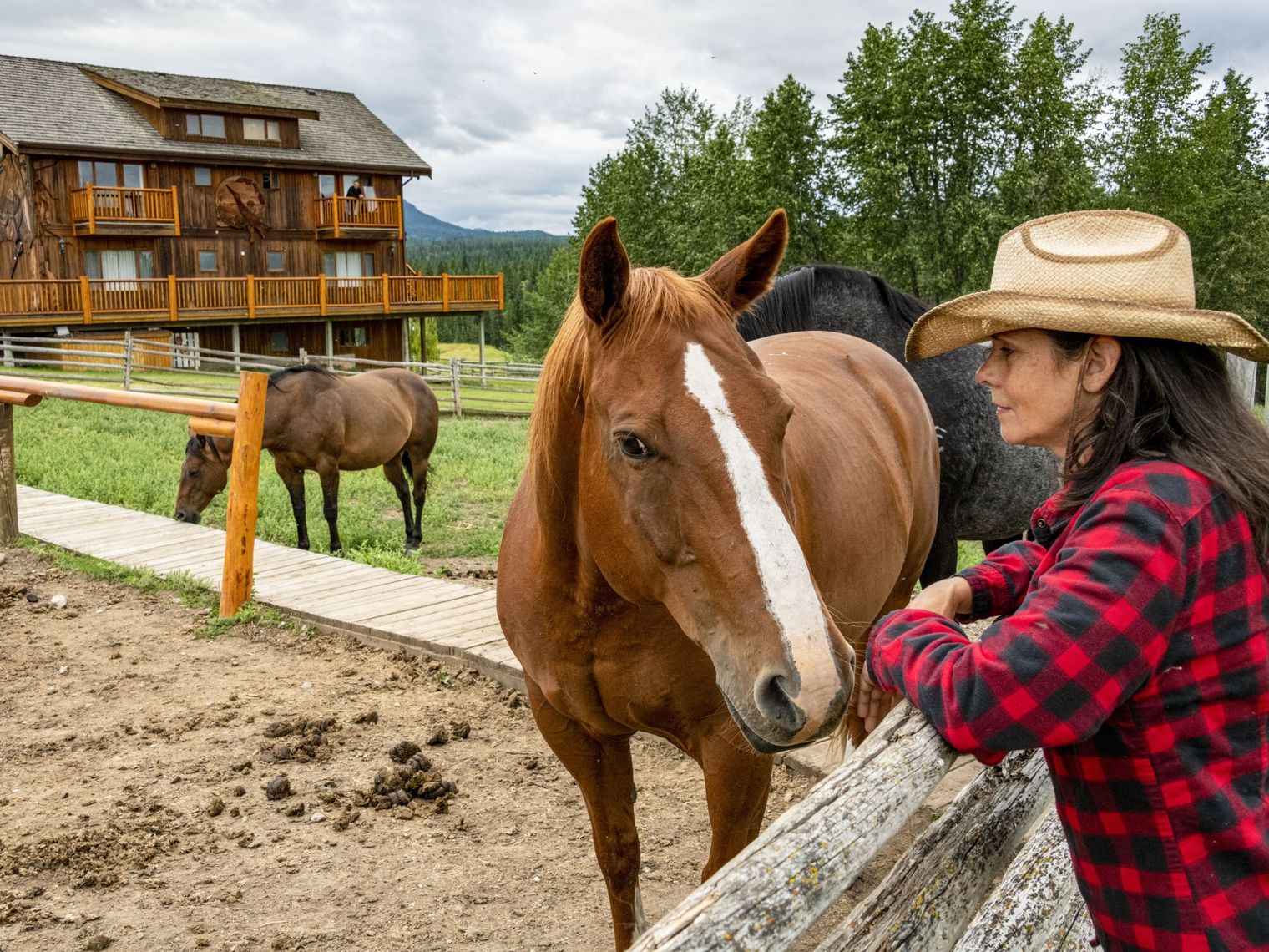 Eine Frau lehnt an einem Zaun neben einem braunen Pferd, im Hintergrund ist eine traditionelle Ranch in Chilcotin, Kanada, zu sehen