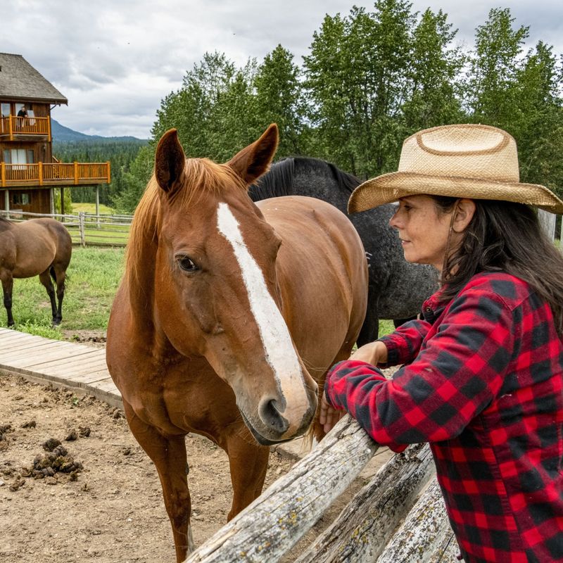 Een vrouw leunt tegen een hek naast een bruin paard, met op de achtergrond een traditionele ranch in Chilcotin, Canada