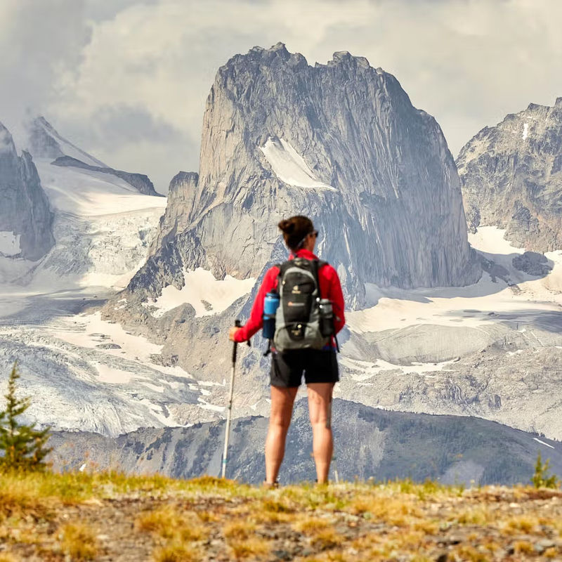 A woman stands on a ridge looking out at the dramatic peaks of the Canadian Rockies, while holding a hiking pole and wearing a backpack
