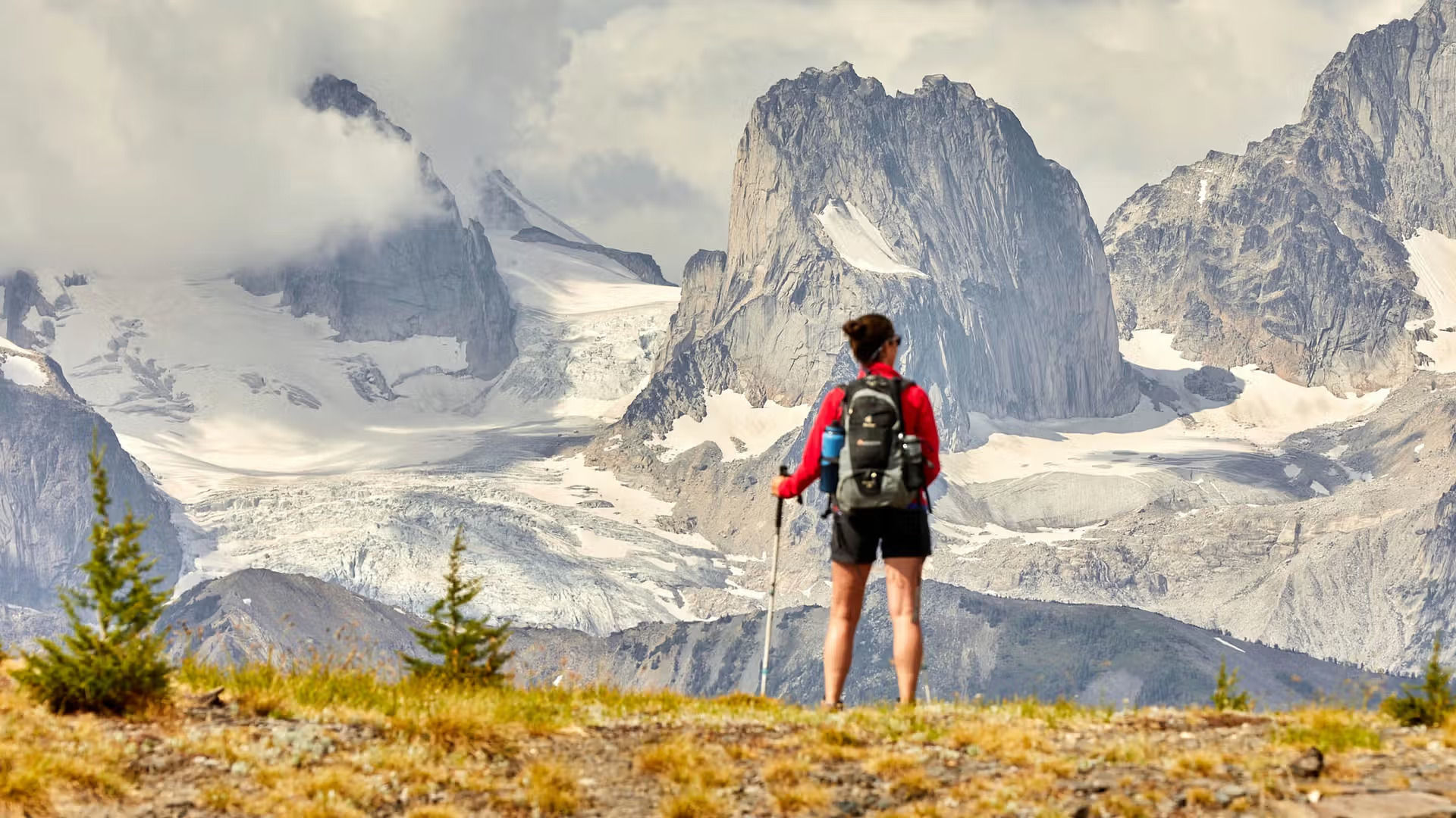 A woman stands on a ridge looking out at the dramatic peaks of the Canadian Rockies, while holding a hiking pole and wearing a backpack