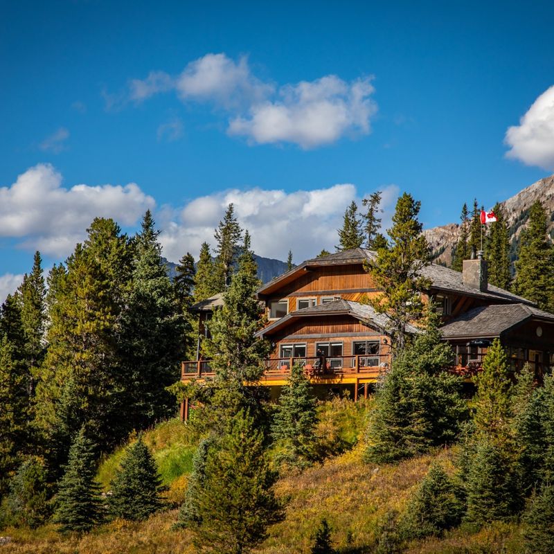 Houten berghut met rondomlopend terras, genesteld tussen sparrenbomen en herfstbladeren in Kananaskis Country, Alberta, Canada, met rotsachtige bergtoppen en een Canadese vlag op de achtergrond.