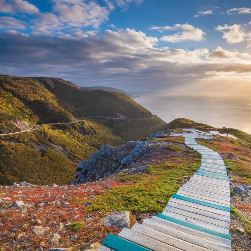 A wooden path winds down the side of a steep cliff on Cape Breton Island, Nova Scotia, with the ocean and sunset in the background