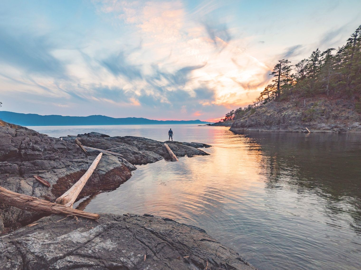 A man standing at the edge of the water looking out towards the sunset and mountains, Sunshine Coast