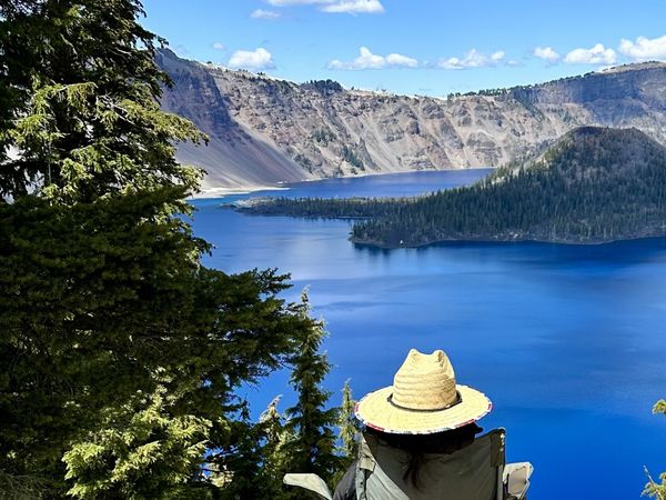 Een vrouw zit in een stoel met uitzicht op een meer in Crater Lake National Park. In het midden ligt een eiland en het beeld wordt omlijst door bomen.