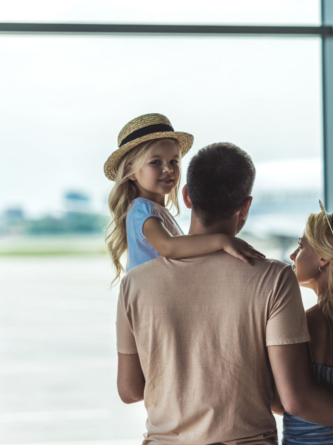 Eine Familie mit zwei kleinen Kindern, die aus dem Fenster eines Flughafenterminals auf ein Flugzeug auf dem Rollfeld blickt, das kurz vor dem Abflug in den Urlaub steht